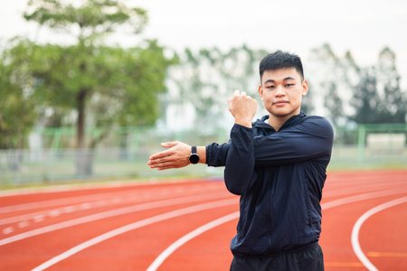 Young asian man wearing sportswear warming up front of sport stadium. Chinese man stretching arms before running on track exercise in the morning. Healthy and athlete lifestyle concept.の写真素材