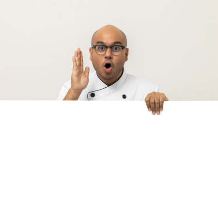 Happy Young handsome asian man chef in uniform looks out from behind an empty whiteboard. Cooking indian man hiding behind big blank billboard for advertising text menu in kitchen and restaurant.の写真素材