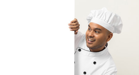 Happy Young handsome asian man chef in uniform looks out from behind an empty whiteboard. Cooking indian man hiding behind big blank billboard for advertising text menu in kitchen and restaurant.の写真素材