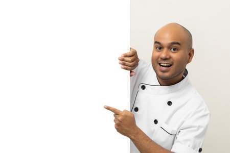 Happy Young handsome asian man chef in uniform looks out from behind an empty whiteboard. Cooking indian man hiding behind big blank billboard for advertising text menu in kitchen and restaurant.の写真素材
