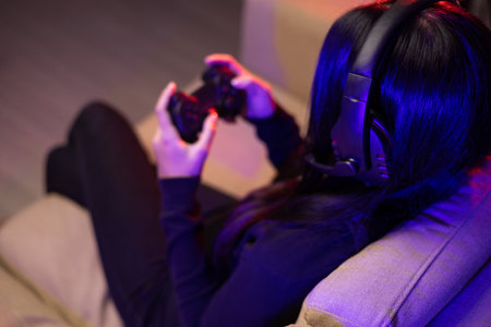 Close up hands Playing video game. Young pretty woman sitting on sofa holding joystick in living room. Female Professional Streamer  wearing hoodie playing game online in dark room neon light.の写真素材