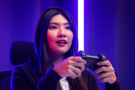 Playing video game. Young asian pretty woman sitting on chair holding joystick in living room. Happy female Professional Streamer chinese wearing hoodie playing game online in dark room neon light.の写真素材