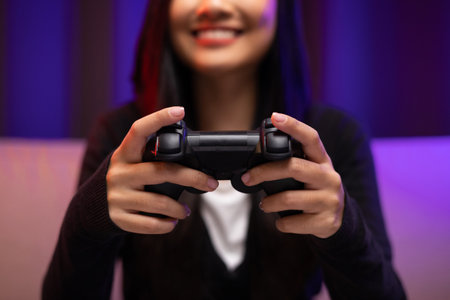 Close up hands Playing video game. Young pretty woman sitting on sofa holding joystick in living room. Female Professional Streamer  wearing hoodie playing game online in dark room neon light.の写真素材