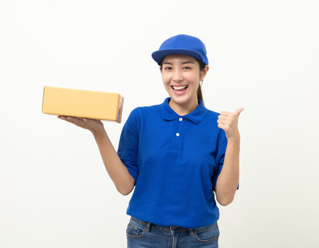 Happy delivery asian woman in blue uniform standing holding parcel cardboard box on isolated white background. Smiling female delivery sending box package service worker.の写真素材