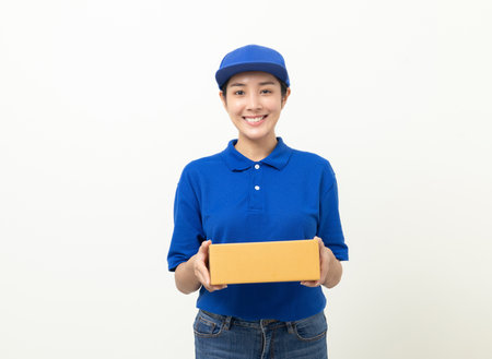 Happy delivery asian woman in blue uniform standing holding parcel cardboard box on isolated white background. Smiling female delivery sending box package service worker.の写真素材