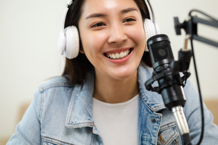 Content creator woman host streaming her a podcast on laptop with headphones and microphone interview cheering guest conversation at broadcast studio. Blogger motivation recording voice over radio.の写真素材