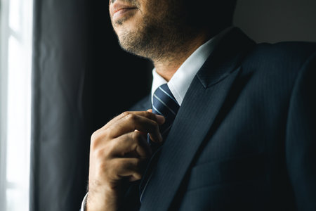 Confident businessman adjust classic blue suit with necktie near window in hotel room. Handsome man wearing a nice suit on wedding day.の写真素材