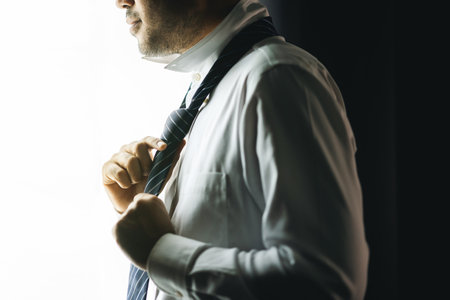 Confident businessman tying or adjust the necktie near window in hotel room in the morning. Handsome man wearing a nice necktie on wedding day.の写真素材
