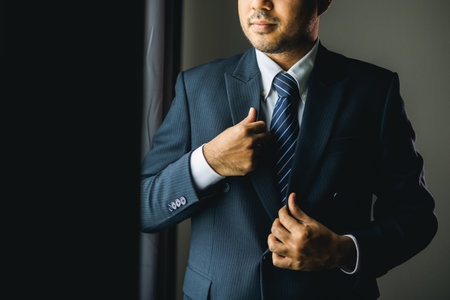Confident businessman buttoning or adjust classic blue suit near window in hotel room at the morning. Handsome man wearing a nice suit on wedding day.の写真素材