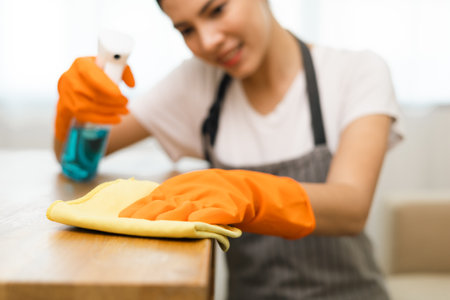 Beautiful housewife with rubber gloves and apron using spray bottle and microfiber towel to clean table at apartment. Young woman is happy to clean home. Maid cleaning service.の写真素材