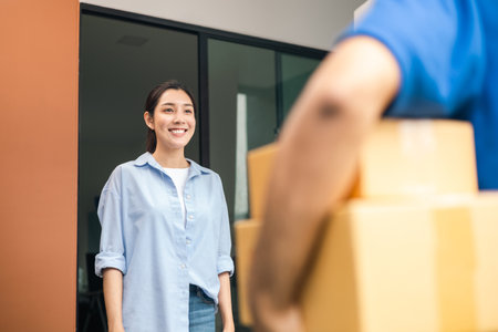 Close up parcel in hand of blue uniform delivery man to customer front of the house with good service from shopping online. Courier man delivering a cardboard box postal package to destination.の写真素材
