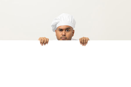 Happy Young handsome asian man chef in uniform looks out from behind an empty whiteboard. Cooking indian man hiding behind big blank billboard for advertising text menu in kitchen and restaurant.の写真素材