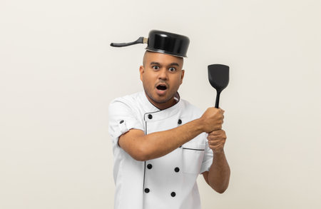 Funny excited face Young asian man chef in uniform put a stew pot over the head holding turner kitchen utensils on isolated. Cooking indian man Occupation chef People in kitchen restaurant and hotel.の写真素材