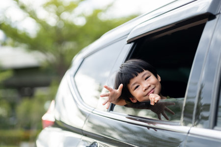 Happy asian boy waving hands gesturing hello out of the car window during a trip with his family....
