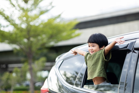 Happy asian boy waving hands gesturing hello out of the car window during a trip with his family. Little child sticking head outta the windshield traveling in a car on a summer vacation.の写真素材