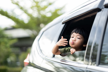 Happy asian boy waving hands gesturing hello out of the car window during a trip with his family. Little child sticking head outta the windshield traveling in a car on a summer vacation.の写真素材