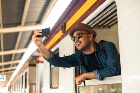 Young asian man travel by train. Sticking his head outta the train window taking photo. Explorer Backpacker arrival and departure at platform railway. Freedom  trip on vacation time holiday weekend.の写真素材