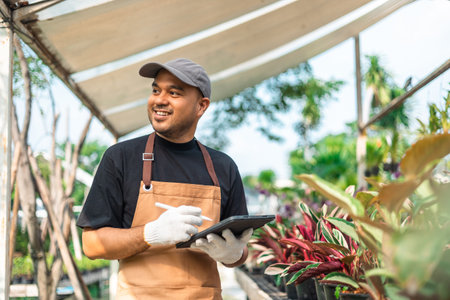 Happy gardener man in gloves plants flowers in greenhouse using tablet check growth quality of Plant. Florists man working gardening in the backyard. Flower care harvesting.の写真素材