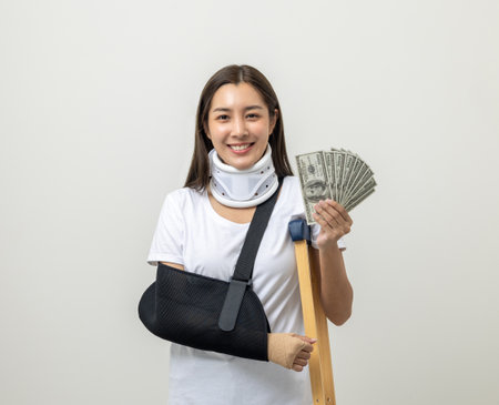Happy young asian woman broken arm holding Dollar banknote. Woman put on plaster bandage cast splint. Patient wearing sling support arm. life insurance and accident have money to spend while sickの写真素材