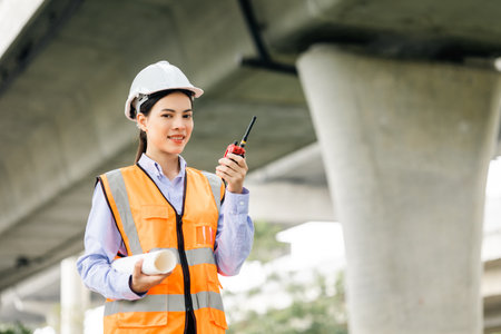 Asian engineer worker woman or architect looking construction with radio wear white safety helmet in construction site. Standing at highway concrete road site. Progress planning of highway bridge.の写真素材