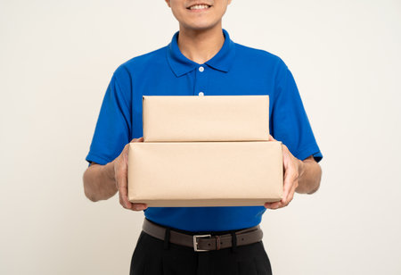Happy delivery asian man in blue uniform standing holding box parcel cardboard on isolated white...