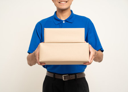 Happy delivery asian man in blue uniform standing holding box parcel cardboard on isolated white background. Smiling male service worker. Delivery courier and shipping service.の写真素材