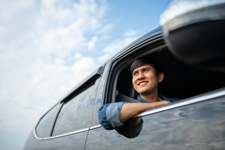 A young Asian man drives a car on a clear day. With beautiful blue sky. He smiling driving to travel by car. Sticking her head outta the windshieldの写真素材