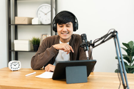 Young asian man host streaming podcast with condenser microphone work on tablet at small broadcast home studio. Content creator blogger recording voice over radio interview guest conversationの写真素材