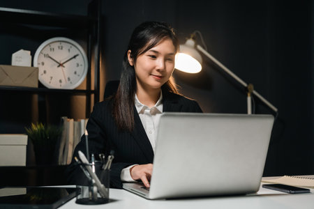 Lovely asian business woman working with laptop at night. Attractive female in suit work late at night overtime sitting in the dark room office.の写真素材