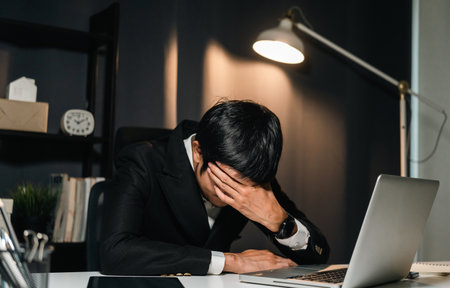 Asian businessman working with laptop computer in the dark office at night. Indian man work hard overtime serious thinking hand on head. Feeling stressed and headache in warm light late at nightの写真素材