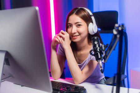 Professional gamer. Young asian pretty woman sitting on chair with computer pc in living room. Happy female Professional Streamer chinese wearing headphone playing game online in dark room neon lightの写真素材