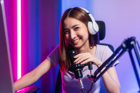 Professional gamer. Young asian pretty woman sitting on chair with computer pc in living room. Happy female Professional Streamer chinese wearing headphone playing game online in dark room neon lightの写真素材