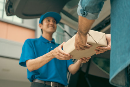 Asian delivery man with parcel in hand of blue uniform sending parcel to customer front of the house from shopping online with good service. Courier man send a package to destination.の写真素材