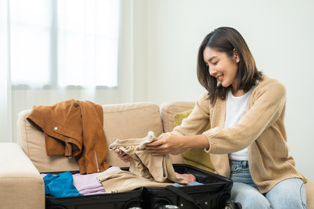 Happy Beautiful woman to folding her clothes pack suitcase for travel in holiday. Young female folding clothes while packing luggage at home.の写真素材