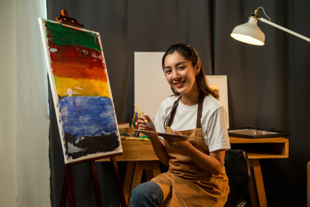 Asian woman artist in the darkroom warm light working on painting with brush and variant acrylic color. Female artist painter on canvas in creative studio as art conceptの写真素材