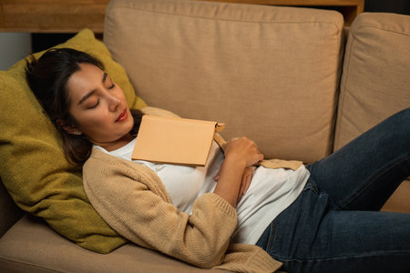 Sleeping Asian beautiful woman lay down on cozey couch sofa while reading book in living room with warm light at night. People female reading book until fall asleep stay home part time relax at homeの写真素材