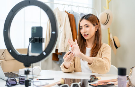 Young beautiful asian woman vlogger live broadcasting cosmetic makeup tutorial content with lipstick looking at cell phone. Influencers with various cosmetic recording review video sharing on socialの写真素材