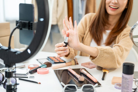 Young beautiful asian woman vlogger live broadcasting cosmetic makeup tutorial content with lipstick looking at cell phone. Influencers with various cosmetic recording review video sharing on socialの写真素材
