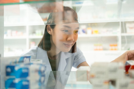 Beautiful asian woman pharmacist checks inventory of medicine in pharmacy drugstore. Professional Female Pharmacist wearing uniform standing near drugs shelves counterの写真素材