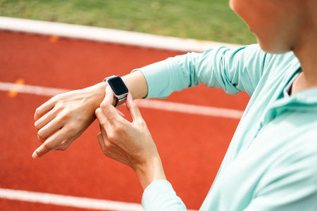 Woman checking sports smart watch on running track at sunrise. Asian female adjusting smart watch before workout outdoor. Exercise in the morning at sport stadium. Healthy and technology lifestyleの写真素材