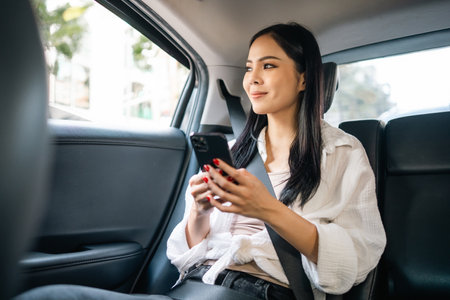 Asian Woman Traveler sitting in car back seats using smartphone call taxi service in application. Online app with cell phone and put on safety belt. Female passenger in car traveling to destination.の写真素材