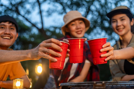 Cheers! Group of asian people friend party camping in nature making toasting with soft drink and beer red cup. Hangout party outdoor in campsite nature forest background on holiday weekend vacationの写真素材