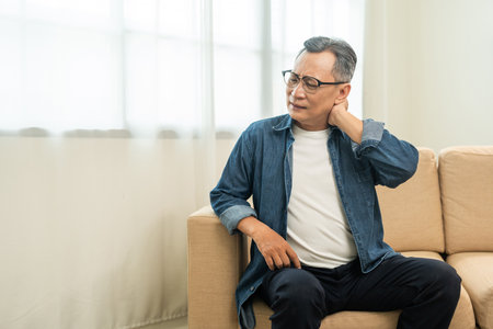 Old man senior has problem with structural posture He had neck and shoulder pain. Mature Massaged his neck and shoulders for relief. reduce muscle tension. Sitting on sofa couch in living roomの写真素材