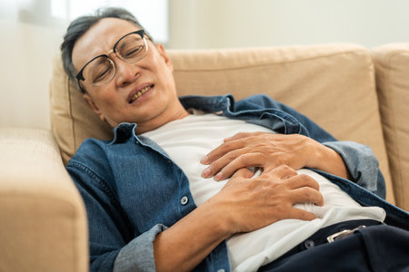 Old man senior had a stomach ache and gastritis. Mature man put hand on his stomach and squeezed it to relax and soothe has go to toilet need to poop. Sitting on sofa in living room at homeの写真素材
