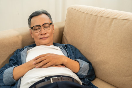 Mature Asian caucasian man lying on comfortable sofa in living room. Asian old man fall asleep take...