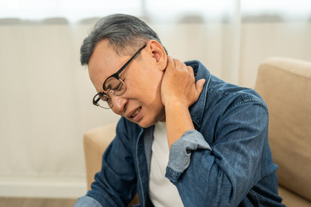 Old man senior has problem with structural posture He had neck and shoulder pain. Mature Massaged his neck and shoulders for relief. reduce muscle tension. Sitting on sofa couch in living roomの写真素材