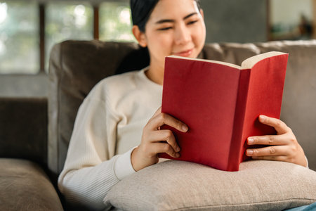 Asian woman sitting reading book on cozey couch sofa in living room with sun light morning. People stay home part time relax at home. Good time quality life rest calm happinessの写真素材