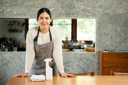 Housewife with apron ready to clean home. Young woman is happy to clean home. Maid cleaning service. Spraying antibacterial sanitizing spray wipe with towelの写真素材