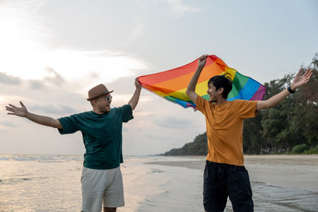 Young couple asian gay with pride movement LGBT holding rainbow flag raise up for freedom. Demonstrate rights LGBTQ celebration pride Month Gay Pride Symbol. Walking on the sand sea beach with sunsetの写真素材