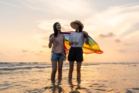Young couple asian lesbian with pride movement LGBT holding rainbow flag for freedom. Demonstrate rights LGBTQ celebration pride Month lesbian Pride Symbol. Walking on the sand sea beach with sunsetの写真素材
